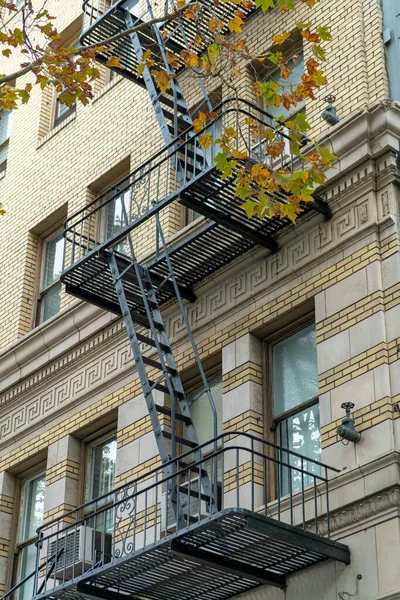 Black fire escape saftey ladder in heart of downtown shady city with gray or beige stucco brick exterior of building. Residential structure in urban area with visible windows and decorative facade.