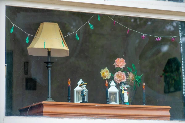 Day of the dead decorations with lamp on table as seen through suburban home or house window in afternoon shade. In city neighborhood in living room for modern mexican holiday designs.