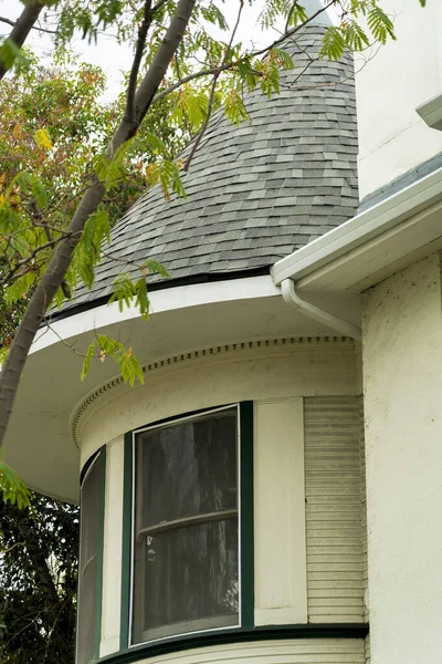 White stucco exterior with turret or spire and gray roof tiles white accent paint on house or home in neighborhood. Late afternoon shade and cloudy sky with back yard trees in secret modern room.
