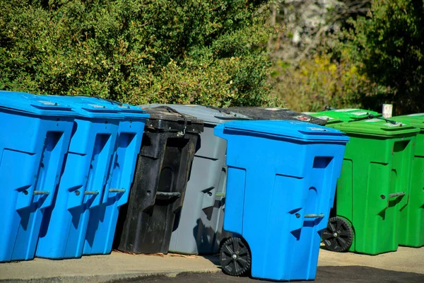 Blue gree black and gray trash cans on street or road for trash day while sanitation workers collect garbage and recycling. In afternoon sun with forest and trees in background neighborhoods downtown.