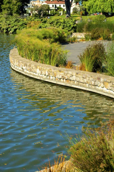 Lakeside cement wall with twisting and winding wall and sidewalk with shrubs and plants on outskirts of pond and water. In afternoon sun in park for feeding ducks and recreation outdoors.