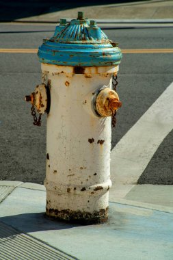 Şehir sokağının köşesinde yaşlanmış ve yıpranmış dış beyaz ve su yeşili rengi olan paslı bir yangın musluğu. Şehrin sanayi bölgesinde beyaz yol boyası ve kaldırımla şehir merkezinde..