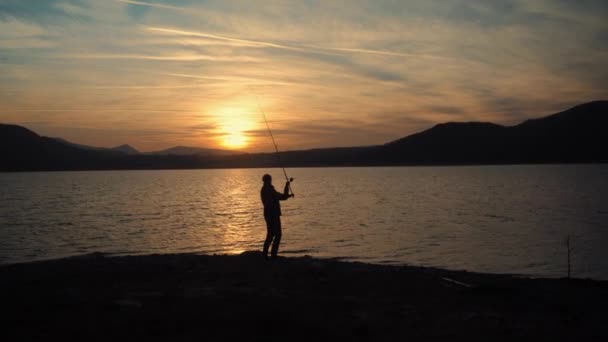 日没の漁師のシルエットは湖にルアーをスローします ロッドで釣る — ストック動画