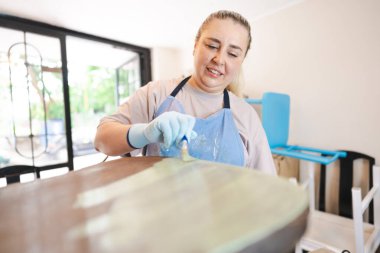 Küçük bir atölyede eski ahşap bir sandalyeyi restore eden, kahverengi gömlek ve eldiven giyen, sandalyeyi boyayan bir kadın. Garajda küçük bir işletme.