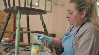 Küçük bir atölyede eski ahşap bir sandalyeyi restore eden, kahverengi gömlek ve eldiven giyen, sandalyeyi boyayan bir kadın. Garajda küçük bir işletme.