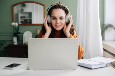 Genç mutlu bayan öğrenci kulaklık takıyor ev bilgisayarında elearning kullanıyor müzik dinliyor, canlı konferans veya podcast webinar dinliyor, online seminerde bilgisayar başında oturuyor.