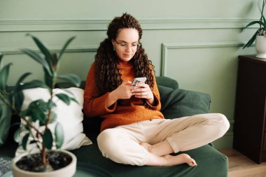 Rahat genç bir kadın evinde elinde akıllı bir telefonla koltukta oturuyor cep telefonlarına bakıyor mobil uygulamalarda alışveriş yapıyor e-ticaret ürünleri sipariş ediyor ya da sosyal medyayı kontrol ediyor.