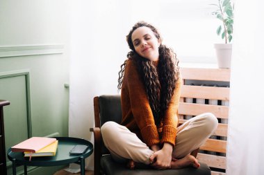 Genç, rahat, gülümseyen, güzel bir kadın evdeki sandalyeye oturmuş dinleniyor. Mutlu, pozitif, güzel bayan mutluluğun tadını çıkarıyor ve modern, rahat bir apartmanın penceresinin yanında dinleniyor.