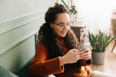 Mutlu genç bir kadın evdeki kanepede online alışveriş için akıllı telefon kullanıyor. Teknolojiden ve e-ticaret için mobil uygulamalardan hoşlanan rahat bir bayan.