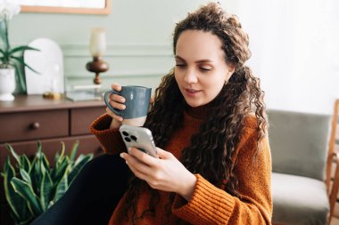Genç bir kadın sıcak bir içkiden ve evde akıllı telefonuyla internetten alışveriş yapmaktan hoşlanıyor. Telefonu ve sıcak içeceği olan mutlu bir bayan, evde e-ticarete dalmış. Pozitif kadın evde rahat bir sabah geçiriyor..