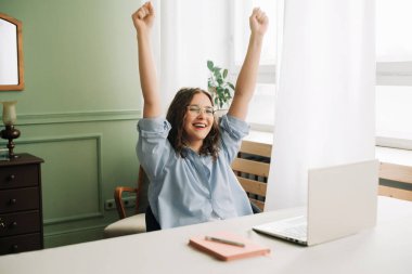 Başarılı genç bir kadın bilgisayarını uzaktan çalışmak ya da çalışmak için kullanırken başarıyı kutluyor. Hedefe ulaşmayı kutluyoruz, çevrimiçi kazanıyoruz, e-posta ile iyi haberler alıyoruz, eller havaya, mutlu hissediyoruz.