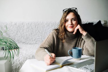 Şık ve odaklanmış bayan halkla ilişkiler müdürü çevrimiçi mağaza için içerik planı oluşturuyor, evde dizüstü bilgisayar, defter ve kahve ile çalışıyor, kameraya bakıyor.