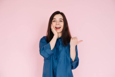 Şaşkınlık ve neşe ifade eden, yanağa dokunan ve pembe izole edilmiş arka planda büyük dişli bir gülümseme ile boş uzayı işaret eden genç bir kadın..