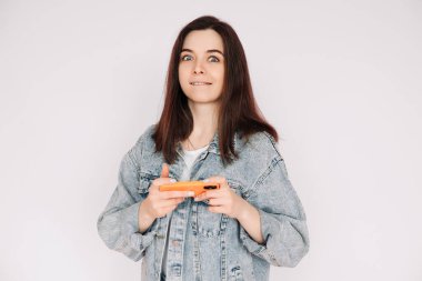 Portrait of a young woman in a casual denim jacket with a smartphone playing a game, isolated against a gray background, conveying indecision and insecurity. clipart