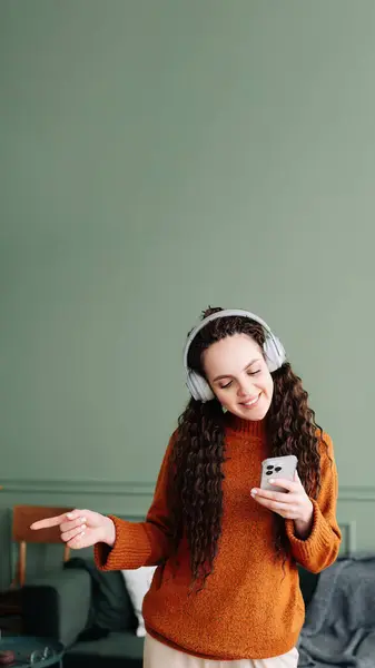 stock image Happy pretty young woman wearing headphones using a smartphone dancing at home. Cheerful girl listening music on mobile phone, singing song feeling relaxed standing in modern living room interior