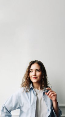 Thoughtful smiling woman with sad eyes in a blue casual shirt poses against the backdrop of a white wall at home, looks away, remembering bittersweet memories of her ex-boyfriend, playing with her clipart