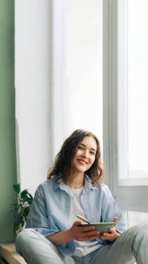 Kot pantolonlu ve pencerede oturan genç bir kadın büyük pencereden içeri bakıyor elinde bir kase kahvaltıyla, gülümsüyor ve dışarıda tuhaf bir şey fark ediyor..