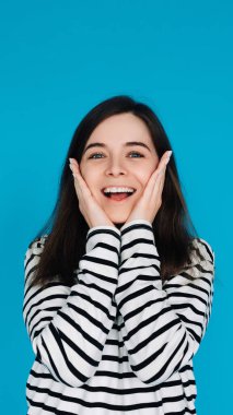 Elmacık kemiklerinde avuç içleri olan, Mavi Arkaplan 'da izole edilmiş, Şaşırmış ve Neşeli Genç Bir Kadının Etkileyici Portresi.