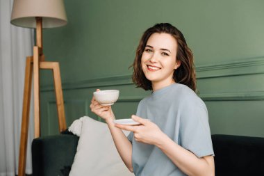 A cheerful pretty young woman with a cup of hot drink sitting on a couch enjoying a vacation. Couch Comfort. Cheerful Woman Savoring Hot Beverage on Holiday. clipart