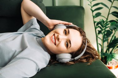 Kablosuz kulaklık takan neşeli genç bir kadın kanepede uzanmış, müzik dinlerken boş zamanlarının tadını çıkarıyor. Müzik mutluluğu. Mutlu Kadın Kablosuz Kulaklıkla Koltukta Dinleniyor.