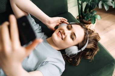 A smiling young middle-aged woman wearing wireless headphones listens to music while lying on the couch with her smartphone. Contentment in Tunes. Smiling Woman Enjoys Music with Wireless Headphones. clipart