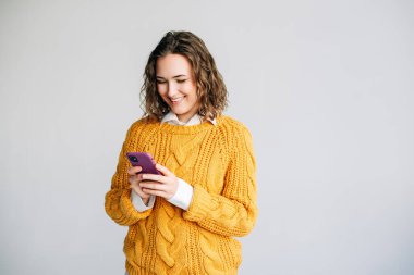 Akıllı Telefon Kullanan Gülümseyen Kadın. Akıllı telefon kullanan kıvırcık saçlı neşeli bir genç kadın. Modern iletişim, teknoloji veya sosyal medya temalarını tasvir etmek için ideal. Beyaz bir arkaplanda izole edilmiş