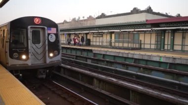Bir metro treni istasyona varır. F hattı işareti olan bir New York metro treni gün batımında istasyona yaklaşır. Yolcular platformda bekleyip, dinamik bir şehir geçişi sahnesi oluşturuyorlar..