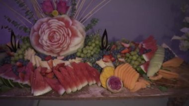 Kavun, kavun, mango, çilek, yaban mersini ve yeşil üzüm gibi çeşitli dilimlenmiş meyvelerle çevrili oyulmuş bir karpuz gülünün sunduğu çarpıcı bir meyve sergisi..