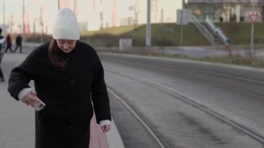 Beyaz şapkalı ve siyah ceketli bir kadın tramvay raylarının yanında dururken akıllı telefonunu kullanıyor. Kentsel ulaşım, toplu taşıma ve modern bağlantı temaları için ideal.