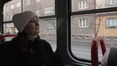 Beyaz şapkalı ve siyah ceketli bir kadın tramvay penceresinin yanında oturuyor ve dışarıdaki binalara bakıyor. Şehir hayatı, toplu taşıma ve düşünceli anlar için ideal..