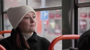 Rahat beyaz şapkalı ve koyu renk ceketli bir kadın tramvay camından dışarı bakıyor, düşüncelere dalmış. Şehirdeki toplu taşıma araçlarına yapılan kış yolculuğunda huzurlu bir yansıma anı yakalıyor..