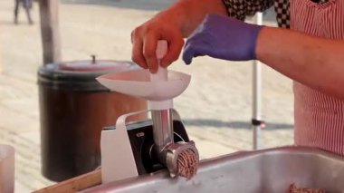 Eldiven ve önlük giymiş bir kasap, açık büfedeki elektrikli öğütücüde çiğ et öğütüyor. Mutfak görselleri, gıda güvenliği temaları veya sokak piyasa içeriği için elverişli