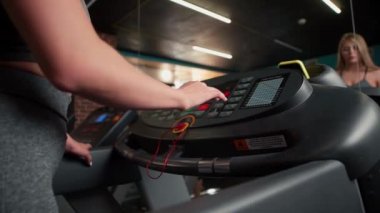 Koşu bandındaki bir spor salonunda bir kadın hareket hızını ayarlamak için düğmelere basar ve simülatörün tırabzanlarını tutarak yürür..