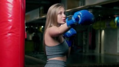 Spor giyimli bir bayan boksör boks yapıyor, bir kız boks eldivenleriyle boks yapıyor, kum torbasının yanında.
