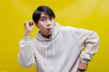 Bej kapüşonlu Asyalı adam mutlu bir şekilde dans ediyor ve sarı arka planda zaferini kutluyor. 