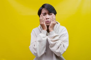 Üzgün bir Asyalı genç adam, elleri ile ağlayan yüz ifadesi sarı bir arka planda izole edilir.