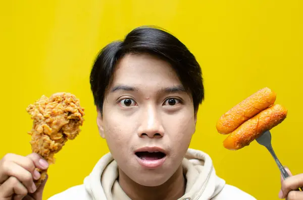 Happy surprised asian man holds fried chicken legs and sausages with wow expression isolated over yellow background. unhealthy fast food concept. food sale promotion