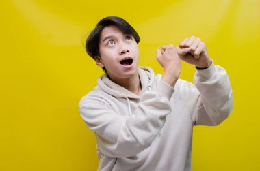 Bej kapüşonlu Asyalı adam mutlu bir şekilde dans ediyor ve sarı arka planda zaferini kutluyor. 