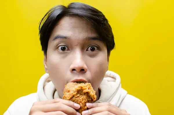 Kendinden geçmiş Asyalı adam sarı arka planda izole edilmiş kızarmış tavuk yer ve ısırır.