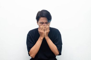Asyalı üzgün genç adamın portresi beyaz arka planda elleri izole bir şekilde yüzünü kapatıyor.   