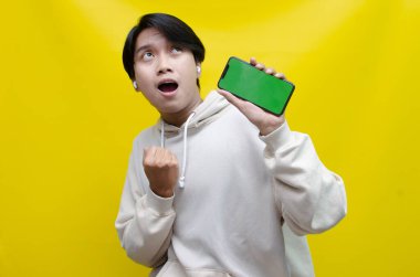 Mutlu Asyalı adam telefonu tutuyor ve yeşil ekran gösteriyor. Asyalı adam bej kapüşonlu yumruğunu kaldırıyor. Ekran telefonunu yeşil ekran şablonuyla en iyi reklam için gösteriyor..