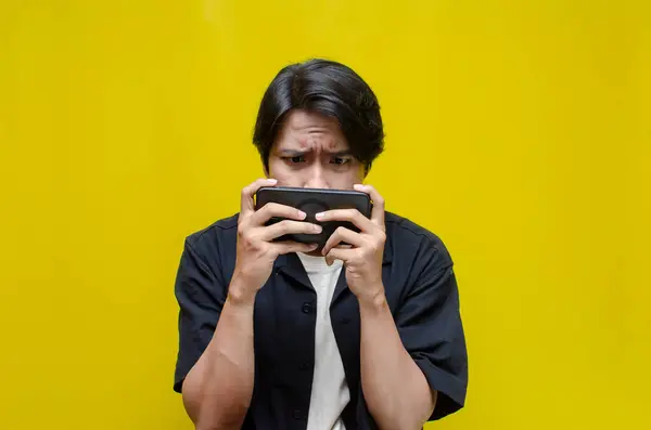 Sarı arka planda cep telefonu kullanan endişeli Asyalı genç adamın portresi   