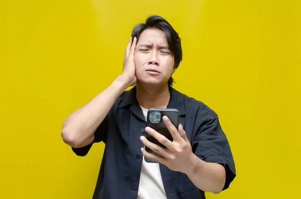 Çaresiz görünen ve cep telefonunu sarı arka planda tutan Asyalı genç adamın portresi.   
