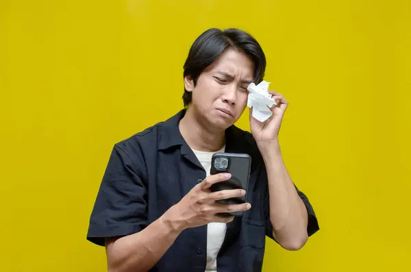 Ağlayan Asyalı adam sarı arka planda akıllı telefonunu kullanıyor. 