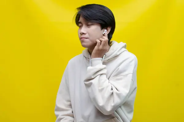 Bej kapüşonlu mutlu, kendini ifade eden Asyalı adam kablosuz kulaklıklarla müzik dinliyor.