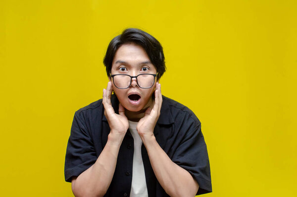 Young Asian man wearing black shirt looking shocked over a yellow background.