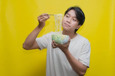 Asyalı Adam, onları çatalla kaldırarak bir kasede erişte yiyor. Krem rengi krem rengi tişörtlü genç Asyalı Çatallı Lezzetli Erişte Çorbası