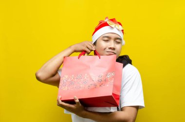 Noel Baba şapkalı genç Asyalı adam sarı arka planda hediye çantaları taşıyor. 