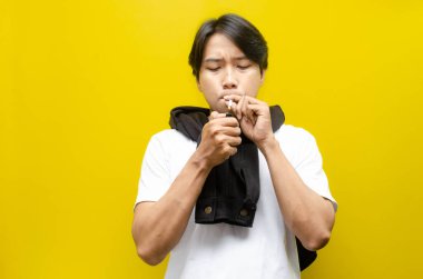 Sarı arka planda sigara içen genç bir Asyalı. Yaşam tarzı seçimini ve sigara bağımlılığı, akciğer hasarı ve genel refah üzerindeki zararlı etkiler de dahil olmak üzere sağlık risklerini vurgular.