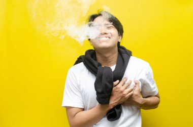 Young Asian man holding his chest in pain while smoking or vaping, with smoke rising. Depicts the health risks of smoking, including chest pain, respiratory issues, and long-term damage clipart